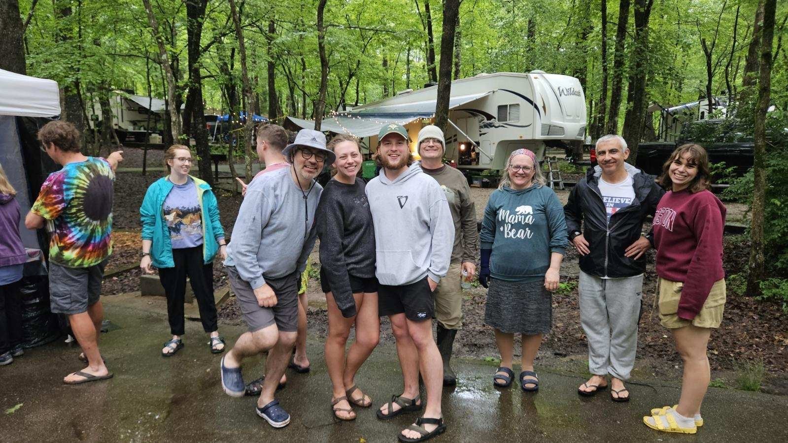 2025 FCF Lara 04 A group of 10-12 people stand in a campsite, some with rain jackets. The ground is wet from the rain. Jonathan looks like he has to go the bathroom because of the way he is holding his legs.