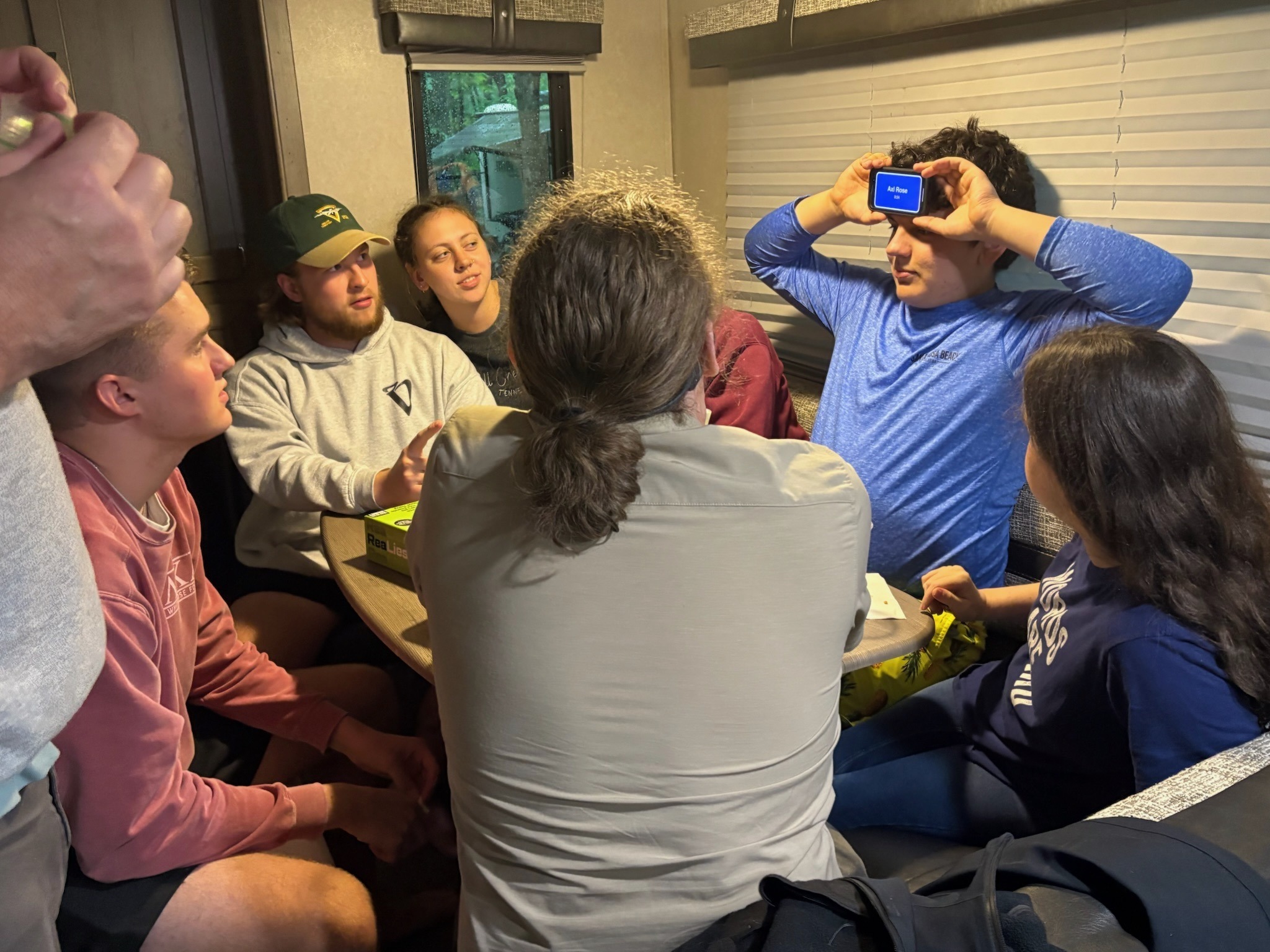 2025 FCF Lara 17 People sitting around a small table inside a camper, playing board games. Caleb is holding a phone to his forehead as part of the game.
