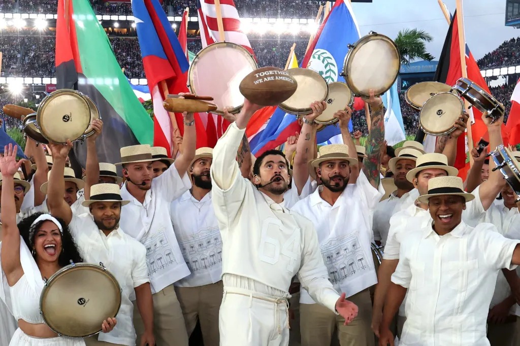 Bad Bunny holds up a football that says "Together We Are America" during his Super Bowl Halftime Show performance in 2026. He is surrounded by Latin men and women holding percussion instruments. In the background are flags of different countries of North and South America.